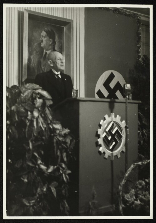 Victor Hämmerle am Rednerpult vor einem Porträt von Adolf Hitler und dem Symbol der Deutschen Arbeitsfront