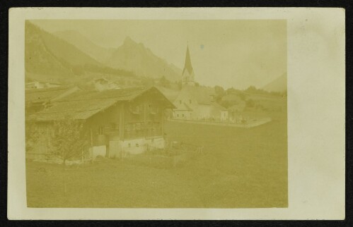 Ortsansicht mit Kirche, Friedhof und Bauernhaus vor Berghintergrund