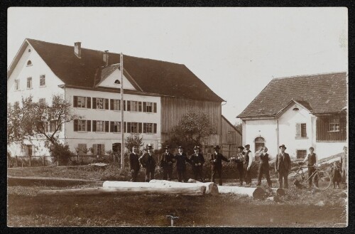 Gruppenbild von elf Männern vor Gebäuden mit Holzstämmen im Vordergrund
